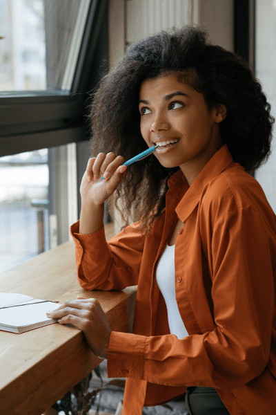 Young Black woman smiles out window holding pen with book on table